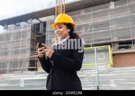 Eine Ingenieurin mit Schutzhelm und formeller Kleidung nutzt auf einer Baustelle ein Smartphone und zeigt die Integration von Technologie in moderne Co-Systeme Stockfoto