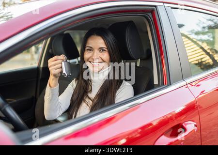 Eine Frau sitzt auf dem Fahrersitz eines roten Autos und lächelt breit, während sie die Autoschlüssel hält. Sie symbolisiert Freiheit und Begeisterung für einen kommenden Straßendreieck Stockfoto
