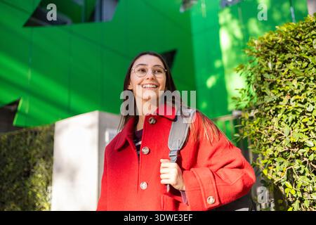 Eine fröhliche Frau in einem leuchtenden roten Mantel lächelt warm und genießt einen hellen Wintertag im Freien mit moderner Architektur und Grün im Hintergrund Stockfoto