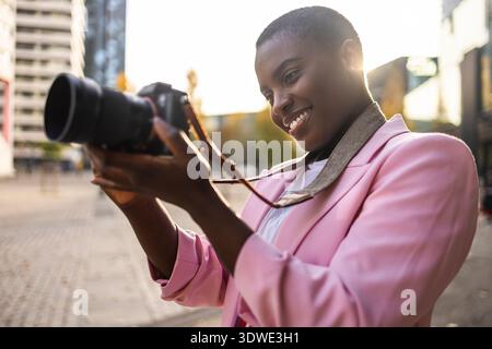 Afrikanische Geschäftsfrau in rosa Blazer lächelt, während sie in einer Stadt draußen eine Kamera benutzt und Momente mit Enthusiasmus und Professionalität festnimmt und zeigt Stockfoto