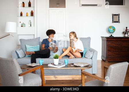 Ein Paar genießt einen entspannenden Nachmittag in seinem gemütlichen Wohnzimmer. Auf einem bequemen grauen Sofa sitzen sie mit einem Tablet und einem Buch und genießen jedes Stockfoto