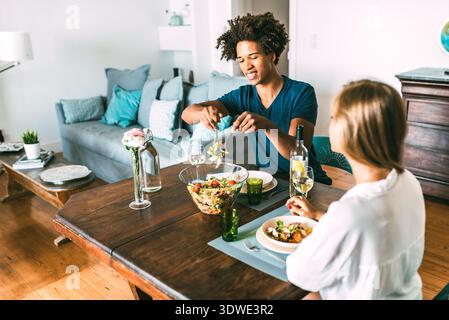 Ein multiethnisches Paar sitzt an einem rustikalen Holztisch in einem gemütlichen Wohnzimmer und teilt eine gesunde Mahlzeit. Sie entspannen sich, während sie die Gesellschaft und d genießen Stockfoto