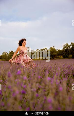 Eine Frau in einem gemusterten Kleid spaziert fröhlich durch ein Lavendelfeld, umgeben von violetten Blüten unter einem bewölkten Himmel. Ihr Ausdruck ist einer von conte Stockfoto