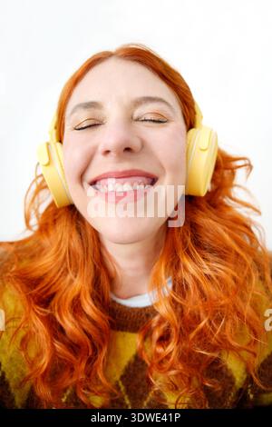 Eine fröhliche Frau mit leuchtend roten Haaren freut sich über ihre Lieblingsmusik. Sie trägt hellgelbe Kopfhörer und drückt Freude und Zufriedenheit aus Stockfoto