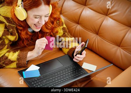 Eine fröhliche Frau mit Kopfhörern liegt auf einer Couch, hält ein Smartphone und eine Kreditkarte. Sie kauft online mit ihrem Laptop ein und genießt den Betrug Stockfoto