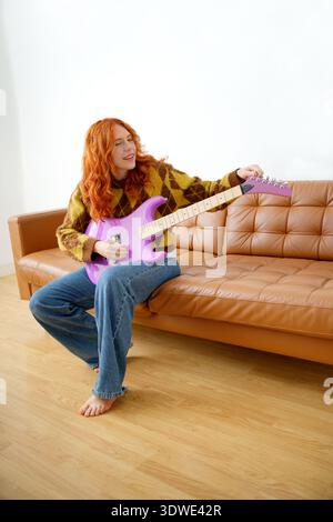 Eine Frau mit rotem Haar sitzt auf einer braunen Ledercouch und lächelt, während sie eine leuchtend violette E-Gitarre tönt. Sie wirkt entspannt in ihrem gemütlichen Hausstuhl Stockfoto