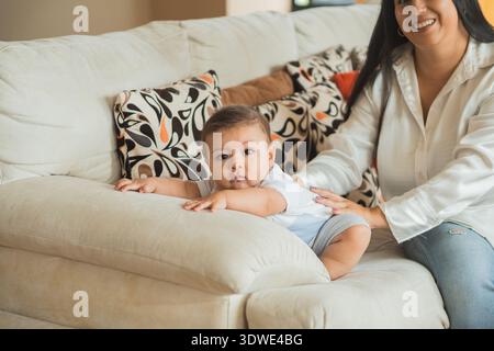 Ein entzückendes Latein-Baby liegt auf einem gemütlichen Sofa, das sanft von einem lächelnden Elternteil gestützt wird. Das Wohnzimmer verfügt über farbenfrohe, gemusterte Kissen, Crea Stockfoto