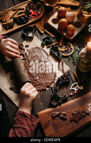 Ein Kind rollt Lebkuchenteig auf einem Holztisch aus, umgeben von verschiedenen festlichen Keksschneidern, Zimtstangen, Sternanis und frischen Eiern und Crea Stockfoto