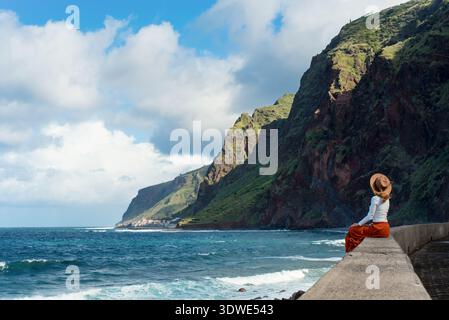 Eine Frau mit Hut sitzt auf einem Steinvorsprung mit Blick auf das Meer und die zerklüfteten Klippen von Jardin do Mar, Madeira, Portugal. Die Szene fängt Ruhe und ein Stockfoto