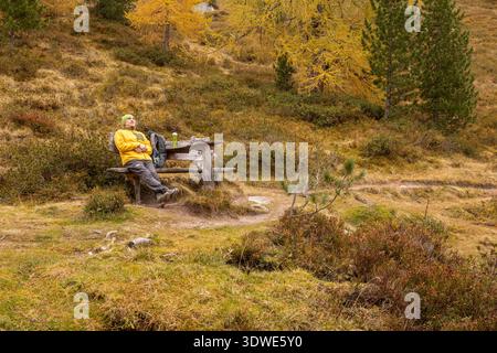 Ein Wanderer in leuchtend gelbem Gang entspannt sich auf einer Holzbank entlang eines malerischen Weges. Goldenes Laub und üppiges Grün sorgen für eine ruhige Szene in den herbstlichen Alpen Stockfoto