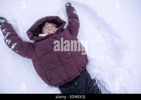 Frau, die fröhlich im Schnee liegt, einen warmen Mantel und eine Brille trägt und einen hellen Wintertag im Freien genießt. Der Schnee bietet eine perfekte Kulisse für eine Stockfoto