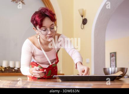 Eine junge Frau mit Brille und festlicher Schürze macht in einer gemütlichen Küche Weihnachtskekse und bereitet sich mit Liebe auf die Weihnachtszeit vor Stockfoto