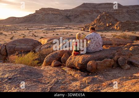 Ein Paar genießt einen ruhigen Sonnenuntergang in Namibias zerklüfteter Landschaft, sitzt auf großen Felsen mit trockenen Ebenen und majestätischen Bergen im Hintergrund, CA Stockfoto