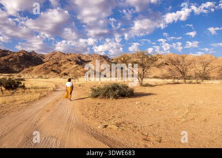 Ein Alleinreisender spaziert entlang eines unbefestigten Pfades in der atemberaubenden afrikanischen Savanne Namibias, umgeben von zerklüfteten Hügeln und verstreuten Bäumen unter einem lebhaften bl Stockfoto