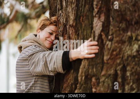 Eine freudige Frau umarmt einen Baum und symbolisiert ihre Verbindung mit der Natur und ihr Engagement für Nachhaltigkeit, umweltfreundliche Praktiken und erneuerbare Ressourcen Stockfoto