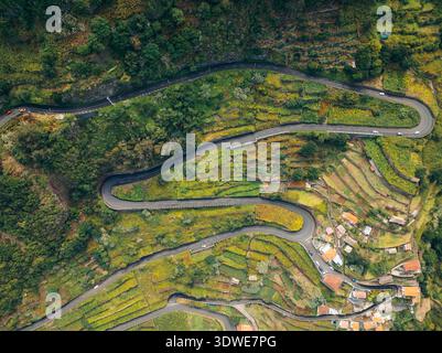 Aus der Vogelperspektive auf eine gewundene Straße durch üppiges Grün und terrassenförmig angelegte Hügel auf Madeira Island, die die lebendige, natürliche Landschaft und das kleine Dorf zeigt Stockfoto