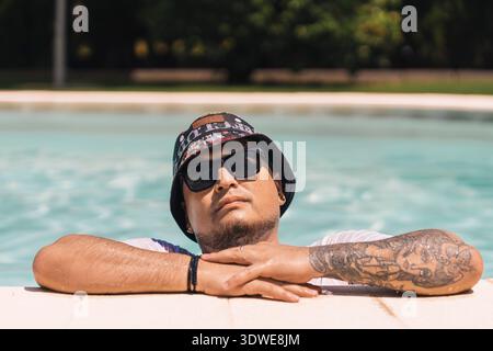 Ein Mann mit Sonnenbrille und Hut genießt einen sonnigen Tag im Pool, während er seine Arme auf dem Rand hält. Die Atmosphäre strahlt ein Gefühl der Entspannung und Pflege aus Stockfoto