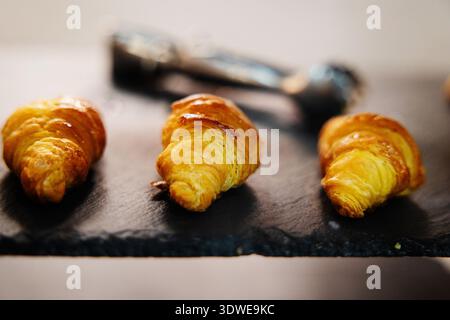 Drei frisch gebackene Croissants sitzen einladend auf einer dunklen Steinplatte. Das goldene, blättrige Gebäck wird vor dem neutralen Hintergrund hervorgehoben Stockfoto