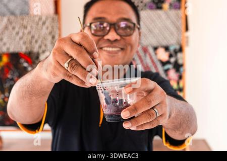 Nahaufnahme eines indonesischen Handwerkers, der einen Plastikbecher mit Batikfarbe hält und den detaillierten Malprozess mit einem feinen Pinsel demonstriert. Traditionelles Ba Stockfoto