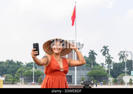 Glückliche Frau, die ein Smartphone hält, einen traditionellen konischen Hut trägt und ein Selfie vor einem Denkmal mit einer vietnamesischen Flagge während der Reise festnimmt Stockfoto