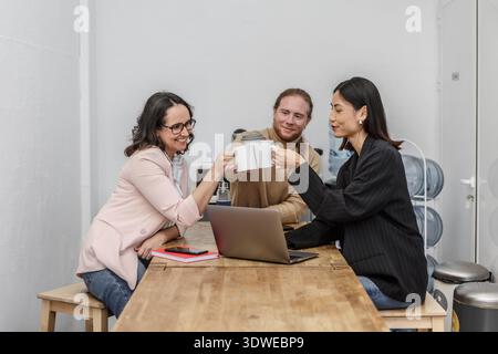 Kollegen treffen sich an einem Holztisch in einem Coworking-Raum, lächeln und toasten mit Kaffeetassen. Ein Laptop und Notebooks sind sichtbar und betonen t Stockfoto