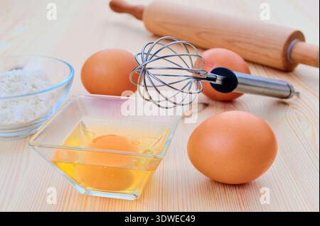 Nahaufnahme von Backutensilien auf einem Holztisch mit frischen Eiern, einer Schüssel Mehl und einem Drahtquirl. Perfekte Zutaten für alle Backzutaten Stockfoto