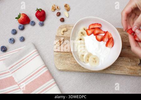 Ein nahrhaftes Frühstück mit einer Schüssel Joghurt mit frisch geschnittenen Bananen und Erdbeeren, umgeben von verschiedenen Beeren und Nüssen auf einem Holz Stockfoto