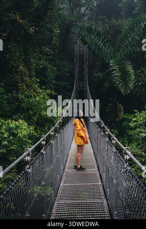 Ein Reisender in einem gelben Regenmantel hält auf einer Metallbrücke im Regenwald Costa Ricas, umgeben von üppigem Grün. Die Szene fängt das Wesen der tr ein Stockfoto