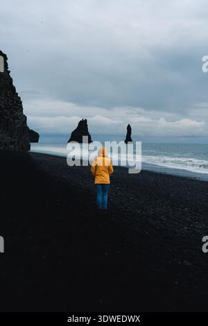 Eine Frau in einer gelben Jacke wandert entlang des atemberaubenden schwarzen Sandstrandes Reynisfjara in Island mit seinen dramatischen Basaltsäulen und Meeresstapeln unter einem Stockfoto