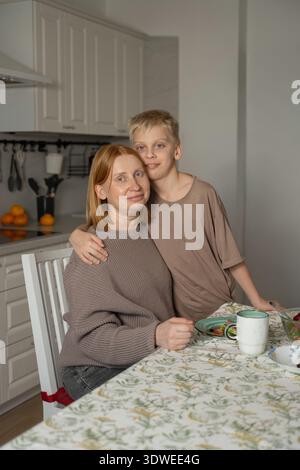 Eine schwangere Mutter und ihr Sohn verbringen einen warmen Moment beim Frühstück in ihrer gemütlichen Küche. Die Einrichtung strahlt wohnlichen Komfort mit einem ordentlich gedeckten Tisch und aus Stockfoto