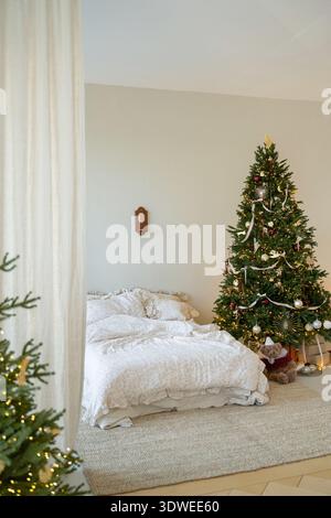 Ein gemütliches, weihnachtlich dekoriertes Schlafzimmer mit einem wunderschön geschmückten Baum mit funkelnden Lichtern und Ornamenten. Das neutrale Bettzeug sorgt für Wärme und Wärme Stockfoto