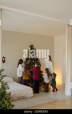 Eine fröhliche Familie versammelt sich um einen wunderschön beleuchteten Weihnachtsbaum, dekoriert ihn mit Ornamenten und Bändern, um die Essenz des Weihnachtsstils und festzuhalten Stockfoto