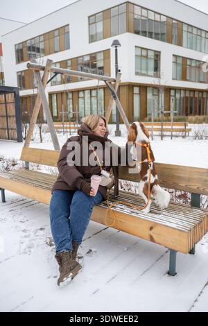 Eine Frau in Winterkleidung sitzt auf einer Bank und genießt einen schneebedeckten Tag mit ihrem Kavalier King Charles Spaniel. Der Hund steht spielerisch auf der Bank, displayi Stockfoto