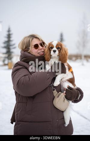 Eine Frau im Wintermantel hält einen Kavalierkönig Charles Spaniel bei einem Spaziergang im Schnee. Die malerische Winterkulisse trägt zum gemütlichen, fröhlichen Ambiente bei Stockfoto