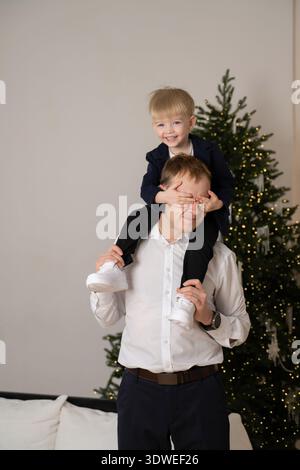 Ein fröhlicher Vater, der seinen lächelnden Sohn auf den Schultern vor einem wunderschön geschmückten Weihnachtsbaum trägt, fängt die Wärme und das Glück des Th ein Stockfoto