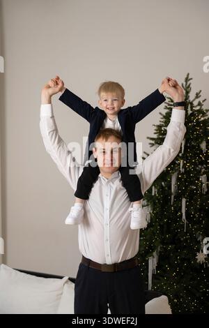 Ein fröhlicher Vater und Sohn feiern gemeinsam die Feiertage. Der Vater hält den Sohn auf den Schultern vor einem wunderschön geschmückten Weihnachtsfest Stockfoto