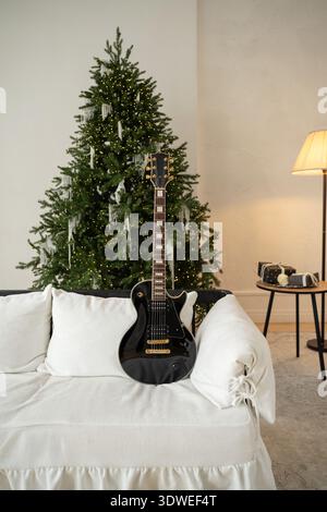 Eine schwarze E-Gitarre sitzt elegant auf einer weißen Couch neben einem wunderschön dekorierten Weihnachtsbaum, mit funkelnden Lichtern für eine warme, festliche Atmosphäre Stockfoto