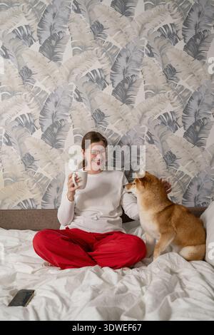 Eine Frau in roter Hose sitzt bequem auf einem Bett und hält eine Tasse, während eine Shiba Inu neben ihr sitzt. Die Umgebung ist hell und gemütlich mit gemusterten Wallpa Stockfoto