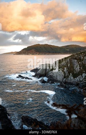 Dramatischer Blick auf den Sonnenuntergang über die felsige Küste bei Cova da Doncela, Praia da Abrela, Lugo, Spanien. Goldene Wolken akzentuieren die Klippen und die Atlantikwellen Stockfoto