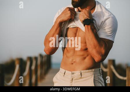 Ein fitter Mann zeigt seinen straffen, muskulösen Körper, indem er sein Hemd hebt, während er auf einem Weg im Freien steht. Das Bild vermittelt einen gesunden Lebensstil und Stockfoto