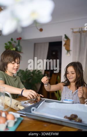 Zwei fröhliche Kinder genießen eine lustige Backsession in der Küche, Formen und bereiten den Teig zusammen mit einem Lächeln zu, umgeben von Zutaten und Backen Stockfoto
