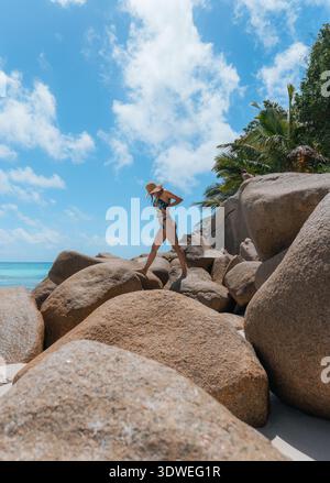 Eine Frau in Badeanzug und Hut erkundet große Felsbrocken am atemberaubenden Strand von Anse Georgette, Praslin, unter einem klaren blauen Himmel mit üppigem Grün in t Stockfoto