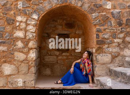 Eine Frau in lebendiger traditioneller Kleidung sitzt friedlich in der Nähe einer antiken Tempelmauer in Indien und spiegelt das kulturelle Erbe und die architektonische Schönheit wider Stockfoto
