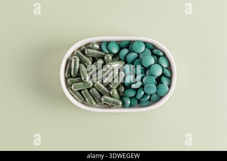 Draufsicht der Spirulina-Kapseln und -Tabletten in einer weißen ovalen Schale auf hellgrünem Hintergrund, die ihre lebendige Farbe und ihren glatten Text unterstreicht Stockfoto