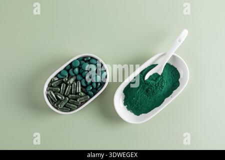 Draufsicht von Spirulina-Tabletten, -Kapseln und -Pulver in weißen Schüsseln auf weichem grünem Hintergrund. Im Pulver liegt ein weißer Löffel, der den vib hervorhebt Stockfoto