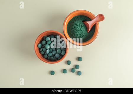 Draufsicht auf Spirulina-Tabletten und grünes Pulver in Terrakotta-Schalen auf einer weichen beigen Oberfläche. Ein Holzlöffel ruht im Pulver, umgeben von Streu Stockfoto
