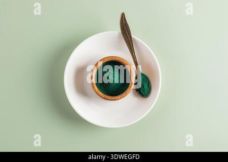 Blick von oben auf Spirulina-Pulver in einer Holzschale und einem Löffel auf einer weißen Keramikplatte mit weichem grünem Hintergrund, die das leuchtende Grün unterstreicht Stockfoto