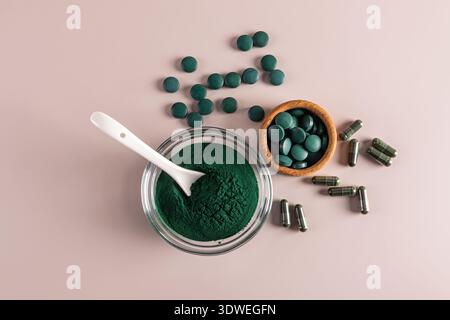 Blick von oben auf Spirulina Pulver in einer Glasschale mit Löffel, umgeben von Spirulina Tabletten und Kapseln. Der Hintergrund ist eine weiche, pastellrosa Oberfläche. Stockfoto