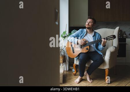 Ein junger Mann sitzt bequem in seinem Wohnzimmer und spielt mit Fokus und Leidenschaft auf der Gitarre. Die gemütliche Umgebung und das Tageslicht sorgen für eine ruhige Atmosphäre Stockfoto