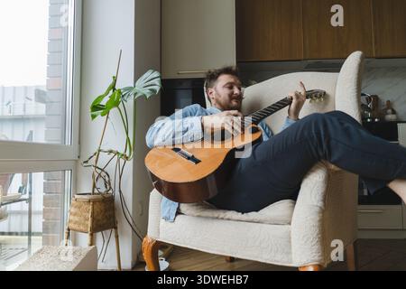 Ein junger Mann entspannt sich zu Hause, sitzt bequem in einem Sessel am Fenster und spielt eine Akustikgitarre. Die gemütliche Umgebung lässt Ruhe und Ruhe vermuten Stockfoto